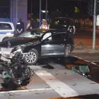 Schwarzes Auto mit schwer beschädigter Front steht nachts auf einer Kreuzung, umgeben von Trümmern und mehreren Personen, darunter Polizisten und Rettungskräfte