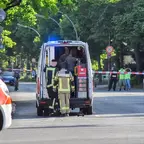 Mehrere Einsatzkräfte in Schutzkleidung stehen an einem geöffneten Einsatzfahrzeug auf einer abgesperrten Straße mit Flatterbändern und Verkehrszeichen im Hintergrund