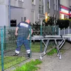 Ein Mann in dunkler Kleidung mit reflektierenden Streifen schiebt nachts eine leere Krankentrage auf einem Gehweg neben einem Zaun in einem Wohngebiet.