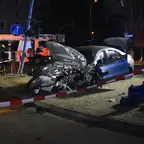 Schwer beschädigtes Auto mit geöffneter Motorhaube an einer abgesperrten Straße in einer nächtlichen städtischen Umgebung.