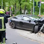 Schwarzes Auto mit stark beschädigter Front prallt gegen einen Ampelmast an einer Straßenecke, drei Feuerwehrleute in gelben Helmen zeigen darauf