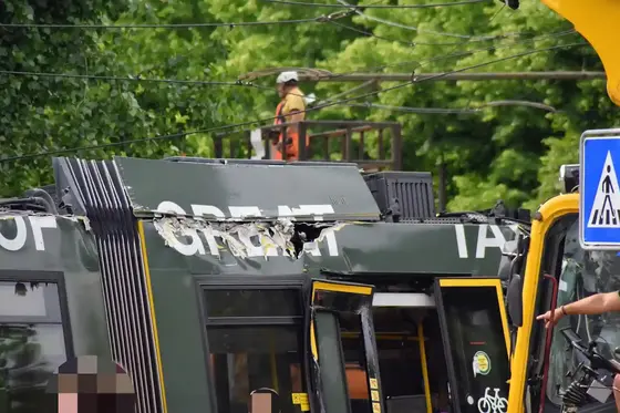 Oben sichtbarer Teil einer beschädigten Straßenbahn mit verbogenem Dach und geöffneten Türen, im Hintergrund ein Bauarbeiter mit Helm auf einer Hebebühne.
