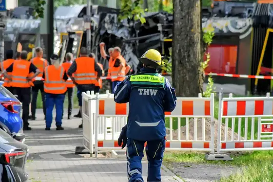 Person in THW-Schutzkleidung und Helm geht auf eine Gruppe von Menschen in orangen Warnwesten zu, im Hintergrund Absperrungen und eine beschädigte Straßenbahn.