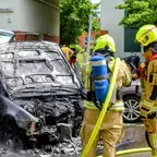 Zwei Feuerwehrleute in Schutzkleidung und Helmen stehen vor einem ausgebrannten PKW mit geöffneter Motorhaube auf einer Straße in Wohngebiet.