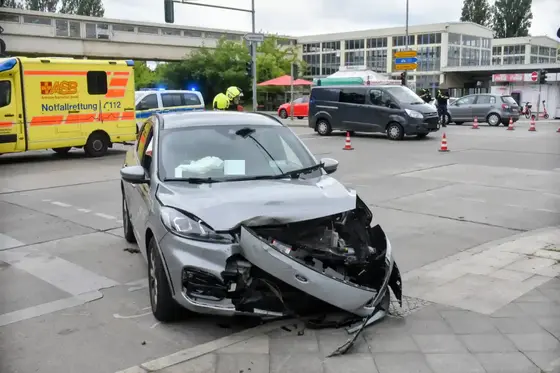 Beschädigter silberner Pkw mit zerstörter Frontpartie steht auf einem Gehweg vor einer Kreuzung mit Einsatzfahrzeugen im Hintergrund.