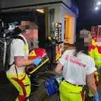Rettungskräfte der Berliner Feuerwehr in gelber Einsatzkleidung und weißem T-Shirt mit Aufschrift 'Berliner Feuerwehr' bei nächtlichem Einsatz vor einem Gebäude mit Notruf-112-Fahrzeug im Hintergrund.