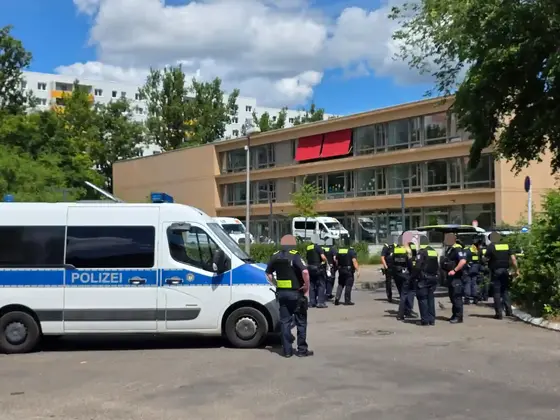Mehrere Polizisten stehen vor einem mehrstöckigen Gebäude mit roten Markisen neben einem weißen Polizeifahrzeug