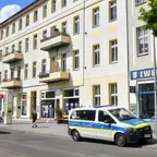 Mehrstöckiges Wohngebäude mit Balkonen an einer Straße, davor ein geparkter Polizeiwagen und mehrere Fußgänger.
