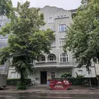 Mehrstöckiges Wohnhaus mit grauer Fassade, umgeben von großen Bäumen, davor ein roter Container und ein weißer Anhänger auf der Straße.