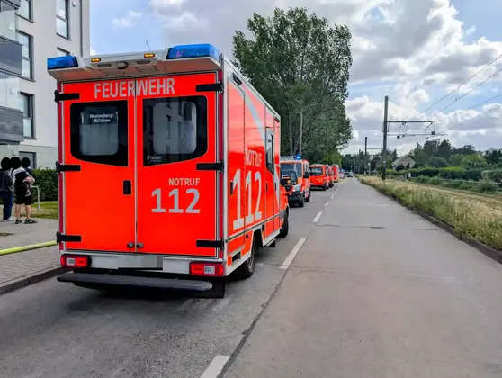 Rote Feuerwehrfahrzeuge mit Blaulicht stehen in einer Reihe an einer Straße neben einem Wohngebäude und einer Straßenbahnstrecke