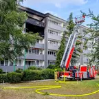 Mehrstöckiges Wohngebäude mit stark verrußtem Balkon im ersten Obergeschoss und ausgefahrener Drehleiter der Feuerwehr davor, Feuerwehrmann steht neben Löschschlauch auf Rasenfläche
