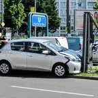 Silberner Kleinwagen steht mit beschädigter Front an einer Werbetafel an einer Straßenkreuzung, im Hintergrund zwei Polizisten und ein Feuerwehrmann in Einsatzkleidung.