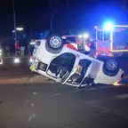 Auf einer Straße liegt nachts ein auf dem Dach liegender weißer Pkw, im Hintergrund stehen Einsatzkräfte und ein Rettungswagen mit Blaulicht.