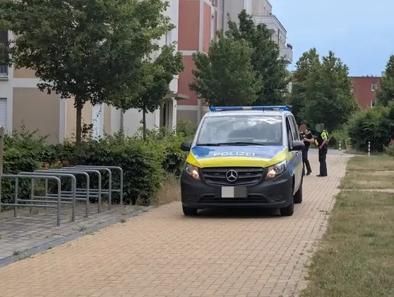Polizeifahrzeug Mercedes-Benz auf gepflastertem Weg vor Wohngebäuden mit zwei Polizisten im Hintergrund.