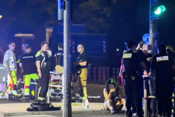 Mehrere Einsatzkräfte der Feuerwehr und Polizei stehen nachts auf einer Straße, eine Person sitzt am Boden und hält sich den Kopf.