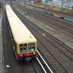 Symbolbild: S-Bahn-Zug fährt auf Gleisen entlang einer mit Graffiti besprühten Mauer in einem urbanen Umfeld.