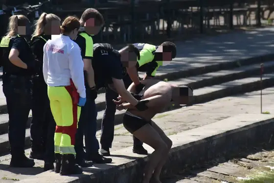Mehrere Polizisten nehmen einen nur mit Badehose bekleideten Mann am Wasserufer fest, eine Person in gelb-roter Rettungsdienstkleidung steht daneben.