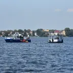 Hausboot und Schiff der Wasserschutzpolizei mit rotem Rettungsboot auf einem See vor Ufer mit Häusern.