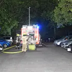 Feuerwehrfahrzeug mit eingeschaltetem Blaulicht und zwei Feuerwehrleuten mit Schutzkleidung auf einer von parkenden Autos gesäumten Straße bei Nacht