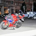 Beschädigtes rotes Motorrad mit sichtbaren Schäden am vorderen Bereich steht auf einer Straße vor einem Gebäude und einem Unfallfahrzeug im Hintergrund.