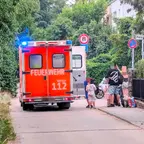 Rückansicht eines roten Feuerwehrfahrzeugs mit Blaulicht auf einer Straße neben einer Gruppe von Kindern und Erwachsenen, die auf dem Gehweg stehen.