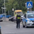 Polizist mit schwarzer Weste und gelber Aufschrift trägt Axt und Armbrust auf einer Straße neben einem Einsatzwagen der Polizei.
