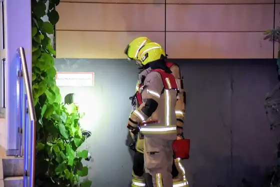 Zwei Feuerwehr-Gruppenführer in Schutzkleidung und gelben Helmen begutachten bei Nacht einen defekten Anschluss an einer Wand, beleuchtet von einer Taschenlampe.