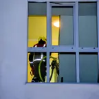 Feuerwehrmann mit Helm und reflektierender Kleidung steht im Treppenhaus hinter einem Fenster, ein Schlauch ist sichtbar.