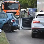 Zwei Autos an einer Unfallstelle auf der Straße, ein blaues Auto mit beschädigtem Heck und offener Fahrertür, dahinter ein Feuerwehrfahrzeug