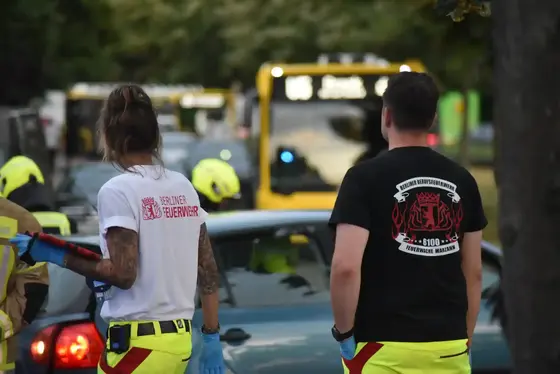 Zwei Personen in gelb-roter Einsatzkleidung stehen auf einer Straße vor wartenden Autos, im Hintergrund ein Bus mit eingeschaltetem Zielanzeiger.