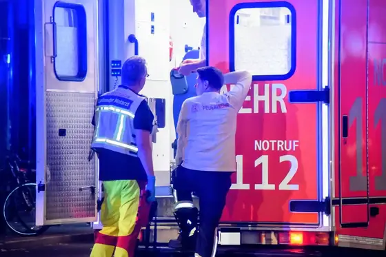 Zwei Einsatzkräfte vor einem geöffneten Rettungswagen, einer trägt eine Weste mit der Aufschrift 'Leitender Notarzt', der andere ein Oberteil mit 'Feuerwehr'