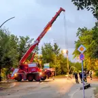 Feuerwehr-Kran hebt eine rechteckige Werbetafel an einer Kreuzung, mehrere Einsatzkräfte stehen daneben, umgeben von Bäumen und Straßenschildern.