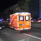 Rückansicht eines Rettungswagens mit eingeschaltetem Blaulicht auf einer nächtlichen Straße neben parkenden Autos und weiteren Einsatzfahrzeugen