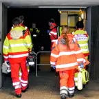 Mehrere Rettungskräfte in orange-roten Schutzanzügen gehen durch einen dunklen Tunnel, eine Person trägt eine gelbe Tasche und eine Sauerstoffflasche.