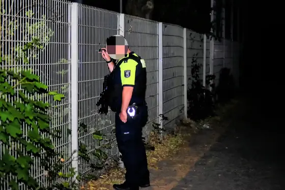 Polizist in Uniform leuchtet nachts mit Taschenlampe durch einen Zaun einer Sportstätte