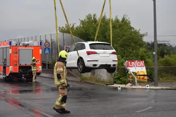 Ein weißes Auto wird mit Endlosschlingen von einem Kran angehoben, Feuerwehrleute in Schutzkleidung stehen auf nasser Straße neben einem Feuerwehrfahrzeug.