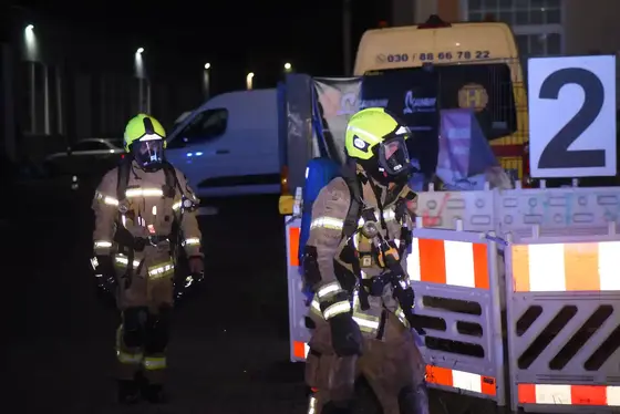 Zwei Feuerwehrleute in Schutzanzügen und Atemschutzmasken gehen nachts an einer Baustellenabsperrung mit der Zahl '2' vorbei.