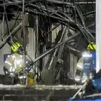 Zwei Feuerwehrleute in Schutzkleidung löschen Glutnester in einem durch Explosion beschädigten Bürogebäude mit freiliegenden Kabeln und zerstörten Wänden.