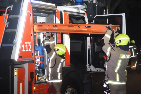 Zwei Feuerwehrleute in Schutzkleidung und Helm heben eine orangefarbene Schleifkorbtrage aus einem geöffneten Feuerwehrfahrzeug mit der Aufschrift 'NOTRUF 112'