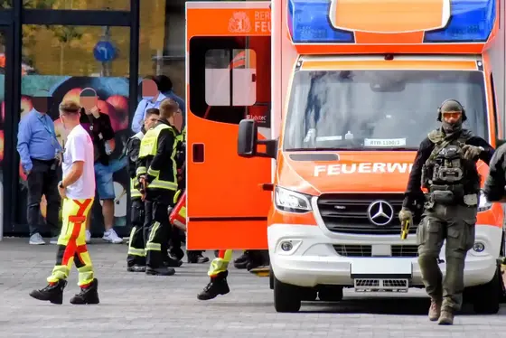 Ein maskierter SEK-Beamter mit Schutzhelm und Distanzelektroimpulsgerät steht vor einem orange-weißen Feuerwehrfahrzeug, im Hintergrund mehrere Rettungskräfte und Personen vor einem Gebäude.