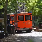Zwei orangefarbene Rettungswagen der Feuerwehr mit der Aufschrift 'NOTRUF 112' stehen auf einem gepflasterten Weg in einem schattigen, von Bäumen gesäumten Bereich, im Hintergrund sitzen mehrere Personen auf einer Bank.