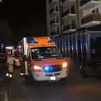 Mehrere Rettungswagen mit Blaulicht stehen nachts in einer Wohnstraße neben geparkten Autos und einem mehrstöckigen Wohngebäude, eine Person in gelber Einsatzkleidung läuft auf der Straße.