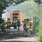 Mehrere Feuerwehrleute in Schutzkleidung gehen auf einem von Bäumen gesäumten Weg auf ein Feuerwehrfahrzeug zu, im Hintergrund ist Rauch zu sehen