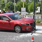 Rotes Auto mit beschädigter Front prallt gegen Geländer einer Haltestelle, umgeben von Glasscherben und Verkehrskegeln, im Hintergrund mehrere Personen und Fahrzeuge.