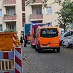 Zwei Fahrzeuge der Berliner Feuerwehr stehen in einer Wohnstraße vor einem mehrstöckigen Wohnhaus, daneben stehen mehrere geparkte Autos und zwei Personen unterhalten sich neben den Fahrzeugen