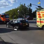 Schwarzes Auto mit Unfallschaden an der Fahrerseite steht auf einer Kreuzung neben einem gelben ASB-Notfallrettungswagen und einem orange-roten Feuerwehrfahrzeug