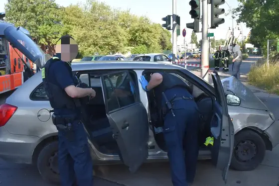 Zwei Polizisten durchsuchen einen grauen Kombi an einer Straßenkreuzung, einer beugt sich in das geöffnete Fahrzeug, der andere steht daneben