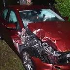 Rotes Auto mit stark beschädigter Front und aufgeblasenen Airbags steht nachts auf Grasfläche neben Straße
