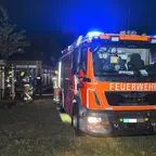 Feuerwehrfahrzeug mit Blaulicht steht nachts auf einer Wiese vor einem Gebäude, Feuerwehrleute in Schutzkleidung sind im Hintergrund aktiv