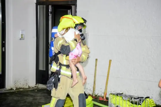 Feuerwehrmann in Schutzkleidung und Atemschutzgerät trägt ein Kind in Nachthemd aus einem Gebäude.